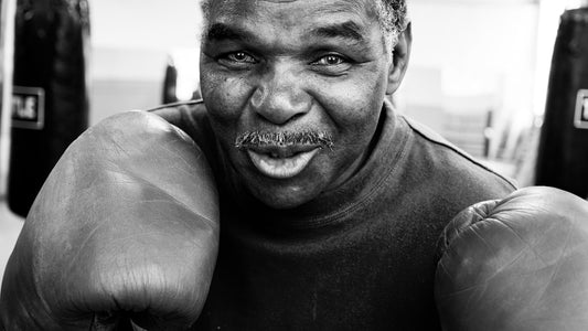 Older boxer smiling in black and white with gloves raised in front of heavy bags