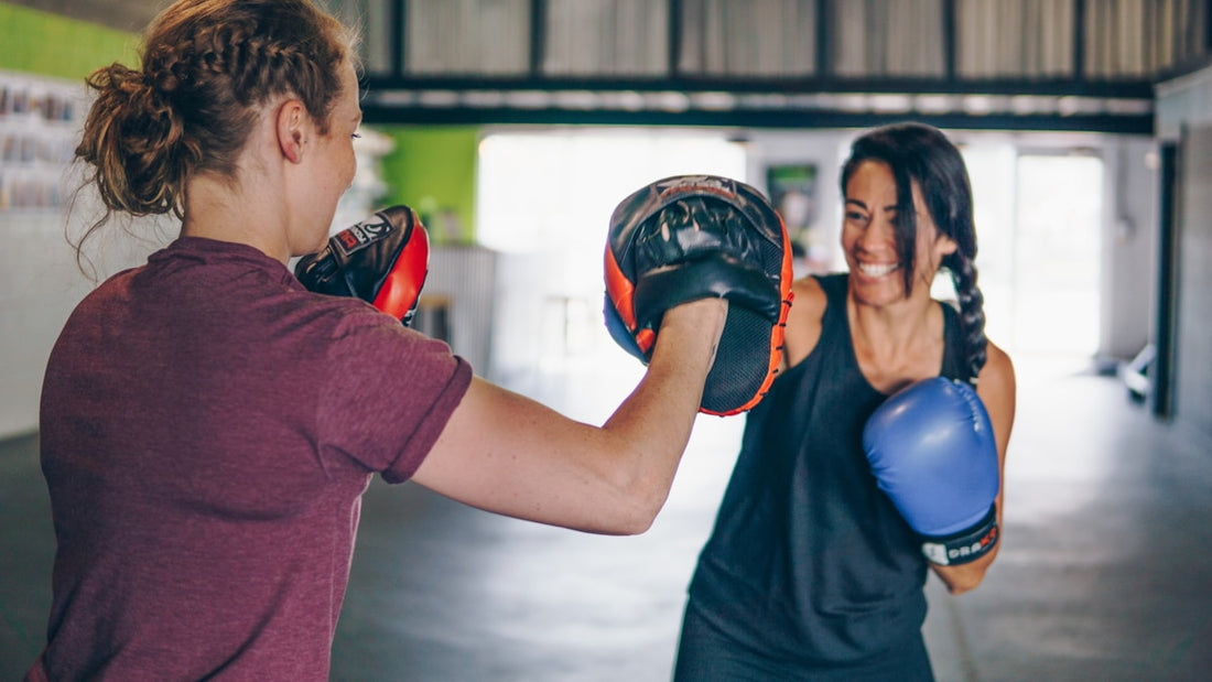 Two women smiling while training together with boxing gloves and focus mitts in a gym